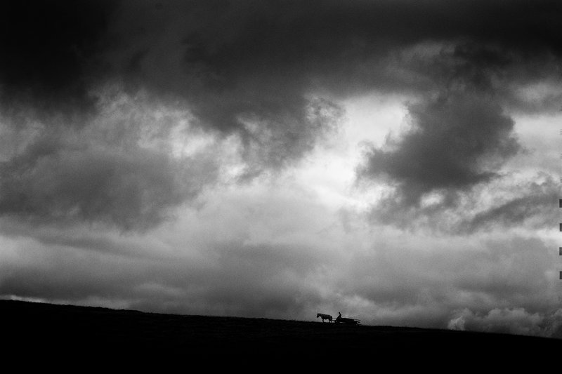 Minimalist black-and-white landscape: dramatic sky with tumultuous dark clouds above a ridge, in the distance a silhouette of a peasant with a horse-drawn cart — a moment of real rural life. Fine-art composition. Awarded photograph on 1x.com.