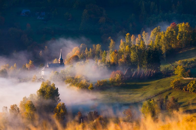 Traditional church with white spire, surrounded by trees, wrapped in waves of low fog in a rural valley at morning. Quiet atmosphere, diffuse light. Awarded photograph on 1x.com.