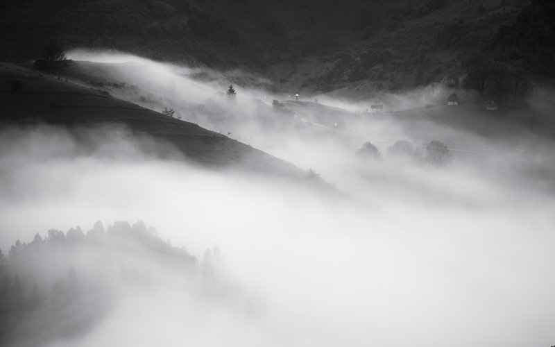 Black-and-white landscape: rolling hills wrapped in waves of dense fog, small traditional houses barely visible among trees. Moody composition, high contrast. Published on 1x.com.