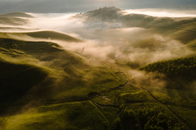 Aerial drone view above a hilly valley with thick golden mist, sun rays piercing through the hills, fresh spring vegetation. Mystical atmosphere, dramatic light. Published on 1x.com.