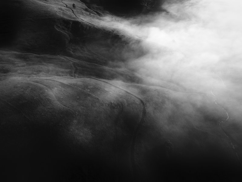 Aerial black-and-white view of rolling hills with winding roads, partially wrapped in bright fog from the right. Abstract composition, high contrast, mysterious atmosphere. Published on 1x.com.