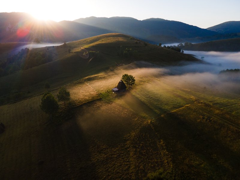 Aerial view at sunrise: lone haystack on a hill with a tree beside it, low waves of fog in the valley, sun rising behind the hills. Pastoral-mystical atmosphere. Awarded photograph on 1x.com.