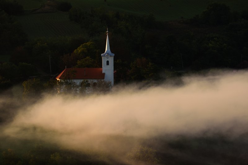 Traditional church with a white spire bathed in warm sunrise light, wrapped by a low wave of fog on the wooded hillside. Picturesque atmosphere, side light. Awarded photograph on 1x.com.