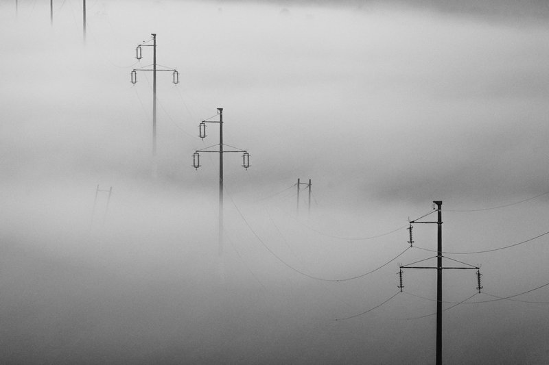 Minimalist black-and-white landscape: high-voltage power poles emerging from a dense sea of fog, connected by barely-visible wires. Abstract industrial-pastoral composition, fine art. Awarded photograph on 1x.com.